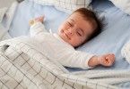 Baby smiling in bed with eyes closed and arms out.