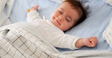 Baby smiling in bed with eyes closed and arms out.